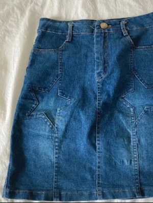 Blå jeanskjol med stjärndetaljer - Säljer en blå jeanskjol med coola stjärndetaljer på framsidan. Kjolen har normal passform, bälteshällor och knappstängning i midjan. Perfekt till vardags för en avslappnad men trendig look.