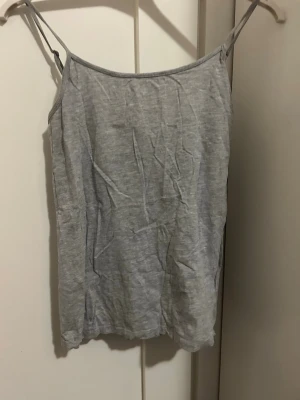 Grå linnetopp från Primark - En enkel och stilren grå linnetopp från Primark med tunna axelband. Toppen är tillverkad i mjuk bomull med stretch för extra komfort. Perfekt att matcha med jeans eller kjol för en avslappnad look. Linnet är i storlek S.