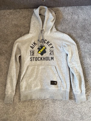 AIK Hockey hoodie - Snygg grå hoodie från AIK Hockey med stort tryck på bröstet där det står 'AIK Hockey 1921 Stockholm' och AIK:s klassiska klubbmärke i svart och gult. Hoodien har huva med dragsko och en stor magficka. Perfekt för dig som vill visa ditt AIK-stöd.
