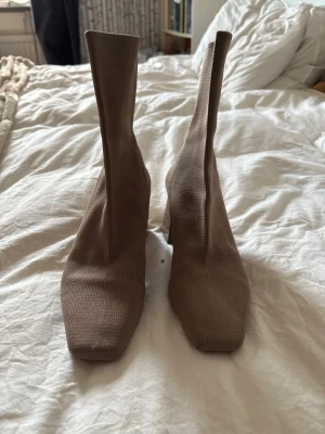 Beige stretchiga stövletter med klack - Säljer ett par beige stövletter med fyrkantig tå och bred klack. Skorna är i ett stretchigt stickat material som sitter tight runt ankeln och ger en modern siluett. Perfekta för dig som gillar trendiga boots med unik form.
