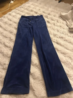 MarinBlå velour mjukisbyxor med vida ben från juicy couture  - Säljer ett par marinblå mjukisbyxor i mjuk velour med breda, utsvängda ben och stora fickor fram. Byxorna har dragsko i midjan och bakfickor med knappdetaljer. Perfekta för en chill och avslappnad stil.