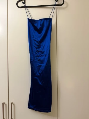 Blå långklänning med tunna band - Snygg blå långklänning i satinliknande material med tunna axelband och enkel, rak siluett. Klänningen har en slits baktill och en elegant, minimalistisk look som passar perfekt till sommarens alla tillfällen.
