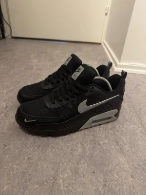 Nike Air Max 90 - Snygga Nike Air Max 90 sneakers i svart med grå detaljer och den klassiska swoosh-loggan i silver reflex material. Skorna har en robust sula med synlig Air Max-dämpning och ovandel i mesh och syntet. Perfekta för dig som gillar streetwear och sportig stil.