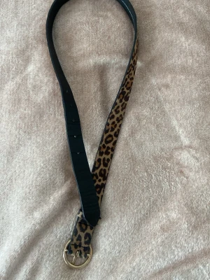 Leopardmönstrat bälte med guldfärgat spänne - Snyggt bälte med leopardmönster och guldfärgat runt spänne. Bältet har en klassisk form och är gjort i skinnimitation. Perfekt för att ge outfiten en cool touch. Den är 92cm lång och storlek xs