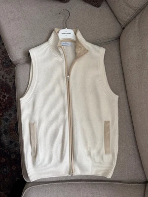 Gran Sasso väst - Säljer en stilren vit stickad väst från Gran Sasso. Västen har ribbad struktur, hög krage och dragkedja framtill. Fickor med beigea detaljer i mockaimitation ger en snygg kontrast. Perfekt att slänga över en skjorta för en clean look.