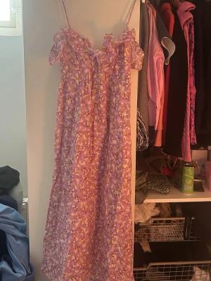 Lång blommig klänning med volang - Söt långklänning med smala axelband och volang vid bysten. Klänningen har ett blommigt mönster i lila, rosa och gula toner. Perfekt för sommarens alla tillfällen och har en luftig, flowy passform.