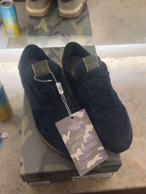 Marinblå sneakers från Valentino - Snygga marinblå sneakers från Valentino Garavani med ovandel i mocka och mesh. Skorna har en klassisk låg silhuett, kamouflagemönstrad detalj på plösen och beige hälparti. Sulan är i gummi och ger bra grepp. Perfekta för dig som vill ha en exklusiv och trendig look. Sulan har släppt men har limmat dit den igen, och skorna generellt är okej skick, lite färg hade släppt men la till lite färg på egen hand 