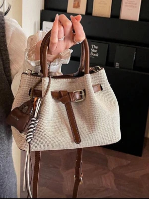 Beige handväska med bruna detaljer - Trendig beige handväska i canvas med bruna läderdetaljer och dekorativt bälte framtill. Väskan har både handtag och justerbar axelrem samt en liten accessoar med nyckelring. Perfekt för dig som gillar stilrena och moderna accessoarer.