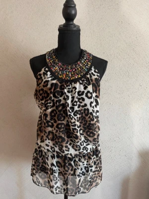 Vintage leopardtopp med stendekoration – ca stl S/M - Den här toppen kombinerar ett klassiskt leopardmönster med en exklusiv halsring dekorerad med färgglada stenar och pärlor.   Detaljer 	•	Storlek: ingen lapp, uppskattas till S/M 	•	Märke: okänt 	•	Mönster: leopard 	•	Halterneck-modell (knyts i nacken) 	•	Dekorativ halsring med många färgade stenar/pärlor 	•	Lätt och följsamt tyg 	•	Ressår i midjan som ger en smickrande passform Skick Fint skick. Inga hål eller fläckar vad jag kan se. Stendekorationen sitter fint.