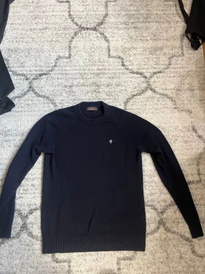 Mörkblå tröja Morris - Mörkblå stickad tröja från Morris med rund halsringning och diskret broderad logga på bröstet. Tröjan har långa ärmar och ribbade muddar vid ärmslut och nederkant. Perfekt för en stilren och klassisk look.