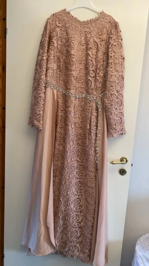  festklänning  - Elegant aprikos färg festgklänning med vackra spetsdetaljer och skimrande bälte i midjan. Klänningen har långärmade ärmar och ett utsvängt fall. Perfekt för dig som vill ha en romantisk och lyxig look.