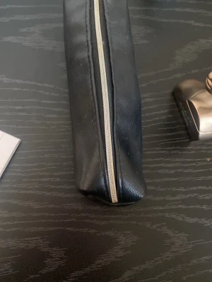 Svart avlång necessär i skinnimitation - En stilren, svart avlång necessär med silverfärgad dragkedja. Tillverkad i skinnimitation och perfekt för att förvara småsaker eller smink. Enkel och minimalistisk design som passar i väskan.