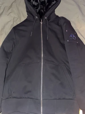 Svart hoodie från Moose Knuckles - Säljer en svart hoodie från Moose Knuckles med dragkedja, huva med snören och mjukt foder. På ärmen sitter det ikoniska metall-logomärket. Hoodien har två fickor framtill och ribbade muddar. Perfekt för dig som gillar streetwear och exklusiva detaljer.