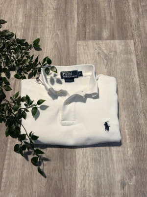 Vit pikétröja från Polo Ralph Lauren - Klassisk vit pikétröja från Polo Ralph Lauren med korta ärmar och krage. Tröjan har två knappar vid halsen och den ikoniska mörkblå logotypen broderad på bröstet. Tillverkad i mjuk bomull som känns skön mot huden.