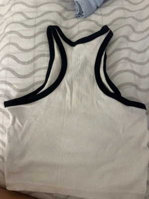 Vit ribbad linnetopp med svarta kanter - Säljer en vit ribbad linnetopp med svarta kontrastkanter. Toppen är ärmlös och croppad med hög halsringning och sportig look. Perfekt att styla med jeans eller kjol för en trendig outfit.