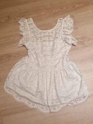 Vit spetsblus med volangärmar - Söt vit blus i spets med rund halsringning och volangdetaljer på ärmarna. Blusen har ett fint mönster och är lätt utsvängd nedtill för en luftig känsla. Perfekt för dig som gillar en ljus, somrig stil.