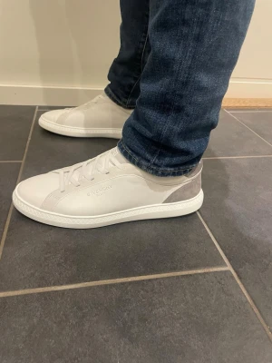 Vita sneakers från Givenchy - Snygga vita sneakers från Givenchy med diskret logga på sidan. Skorna har en låg profil och är tillverkade i slätt läder med grå mockadetalj vid hälen. Skorna är helt nya och oanvända och kommer med box, certifikat och påse! Storlek 44 EU men lite stora i storleken och passar nog även folk med 45!