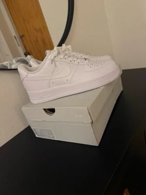 Nike Air Force 1 vita sneakers - Klassiska Nike Air Force 1 sneakers i helvitt läder med låg profil och ikonisk swoosh på sidan. Skorna har perforerad tå för ventilation, vit sula och matchande vita skosnören. Perfekta för en clean och stilren look.