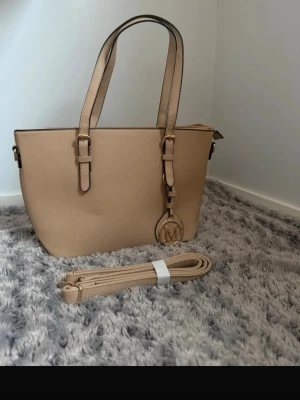 Beige handväska  - Snygg beige handväska i stilren design med två handtag och avtagbar axelrem. Väskan har guldfärgade detaljer, M-hänge och dragkedja upptill. Tillverkad i tåligt konstläder och har en praktisk ytterficka med dragkedja på baksidan.