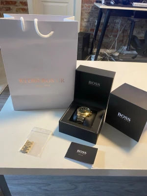 Guldfärgad klocka från BOSS - Stilren guldfärgad klocka från BOSS med metallarmband och grön urtavla. Klockan har kronograf-funktion, datumvisning och detaljer i guld och svart. Levereras i originalask med extra länkar och certifikat.