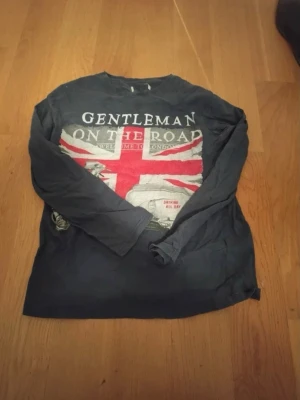 Mörkblå långärmad t-shirt Gentleman - Cool mörkblå långärmad t-shirt med stort tryck av Union Jack och texten 'Gentleman On The Road' på bröstet. Perfekt för dig som gillar brittisk stil och vill sticka ut. Skön passform och mjukt material, passar till jeans eller chinos.