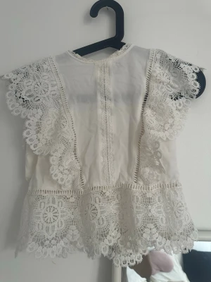Vit spetsblus med korta ärmar - Supersöt vit blus med detaljerad spets och korta ärmar. Blusen har vackra virkade spetsmönster längs ärmar och nederkant samt pärlknappar i ryggen. Perfekt för dig som gillar romantisk och bohemisk stil.