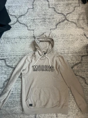 Beige hoodie från Morris - Snygg beige hoodie från Morris med stor logga framtill. Hoodien har vit dragsko i huvan och en klassisk känguruficka. Tillverkad i mjukt bomullsmaterial som känns skönt mot huden. Perfekt för en avslappnad och trendig look.