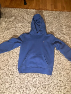 Blå hoodie från Polo Ralph Lauren - Snygg blå hoodie från Polo Ralph Lauren med klassisk huva och magficka. Liten broderad logga på bröstet i vitt. Tillverkad i mjukt material som känns skönt mot huden. Perfekt för en avslappnad och stilren look.