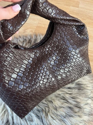 Brun flätad handväska i skinnimitation - Säljer en snygg brun handväska med flätad struktur och mjuk, rundad form. Väskan har ett kort handtag och är tillverkad i skinnimitation med glansig finish. Perfekt accessoar för dig som gillar trendiga detaljer och stilren design.