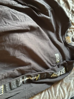 Svart sweatshirt från Emporio Armani - Svart sweatshirt från Emporio Armani EA7 med tryckt logga på bröstet och detaljer längs ena ärmen. Tröjan har rund halsringning och är tillverkad i mjukt bomullsmaterial. Perfekt för en avslappnad och trendig look.