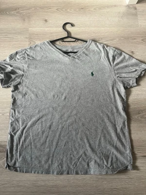 Grå v-ringad t-shirt från Ralph Lauren - Snygg grå t-shirt från Ralph Lauren med v-ringning och klassisk grön broderad logga på bröstet. Tillverkad i mjuk bomull och har korta ärmar. Perfekt basplagg för en clean och enkel stil.