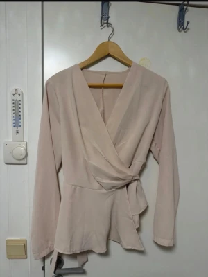 Ljusrosa omlottblus med knytband - Trendig ljusrosa omlottblus med långa ärmar och knytband i midjan. Blusen har en djup v-ringning och är tillverkad i ett mjukt, tunt tyg som ger ett elegant fall. Perfekt för dig som gillar stilrena och feminina plagg.