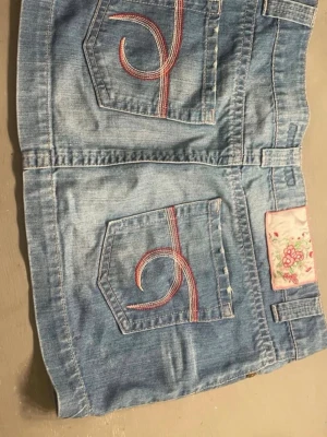 Blå jeanskjol med broderade fickor - Snygg ljusblå jeanskjol i klassisk modell med två bakfickor dekorerade med rosa broderade detaljer. Kjolen har framfickor, bälteshällor och en knappstängning framtill. Materialet är slitstarkt denim och det finns en söt etikett med blommönster bak. Passar mig som har XS super fin till sommaren ,köp gärna 💗💗