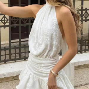 Vit halterneckklänning med spets - Säljer en elegant vit klänning med halterneck och vackra spetsdetaljer. Klänningen har omlottkänsla i midjan och volang nedtill. Perfekt för dig som vill ha en romantisk och stilren look.