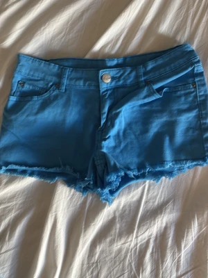 Blå fransiga jeansshorts - Säljer ett par blå jeansshorts med fransig kant och klassisk femficksdesign. Shortsen har knapp och dragkedja framtill samt bälteshällor. 