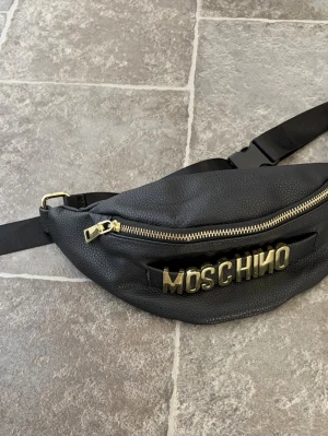 Moschino väska över bröstet eller midjan - Snygg svart från Moschino i strukturerat läder med guldfärgade detaljer och dragkedja. Väskan har ett brett band och ett stort MOSCHINO-logo i metall framtill. Perfekt för dig som vill ha en trendig och praktisk accessoar.