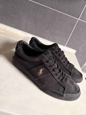 Svarta Polo Ralph Lauren sneakers - Snygga svarta sneakers från Polo Ralph Lauren med klassisk låg profil. Skorna har canvasmaterial, svart yttersula och den ikoniska Polo-loggan broderad på sidan. Perfekta för en clean och stilren look.