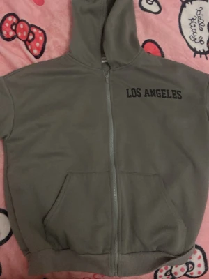 Grå hoodie med tryck Los Angeles - Grå zip-hoodie med svart text 'LOS ANGELES' på bröstet och en stor svart stjärna på ryggen. Hoodien har huva, dragkedja framtill och en stor magficka. Perfekt för en chill och avslappnad stil. Materialet känns mjukt och bekvämt.