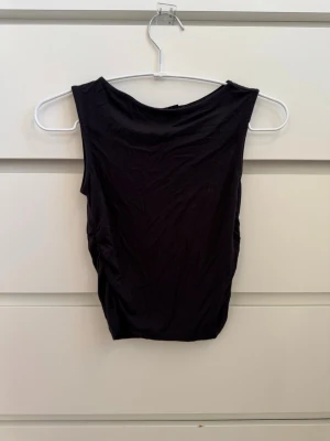 Svart ärmlös linnetopp - Snygg svart ärmlös linnetopp med båt-halsringning. Toppen är väldigt mjuk och har rynkade sidor som gör att den sitter tight och formar sig efter kroppen 😍
