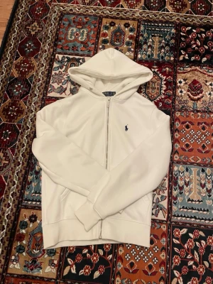 Vit hoodie från Polo Ralph Lauren - Snygg vit hoodie från Polo Ralph Lauren med dragkedja framtill och klassisk blå logga broderad på bröstet. Hoodien har huva och fickor på sidorna. Perfekt för en clean och stilren look.