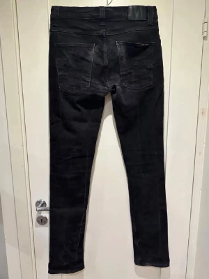 Nudie Grim Tim  - Säljer ett par svarta jeans från Nudie Jeans. Jeansen har klassisk femficksmodell, raka ben och diskret logga på bakfickan. Tillverkade i stretchigt bomullsmaterial för skön passform. Perfekta till en clean och stilren look.
