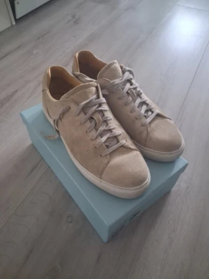 Beige mockasneakers med vit sula - Stilrena sneakers i beige mocka med matchande beige skosnören och vit gummisula. Låg modell med klassisk design och brunt innerfoder. Perfekta för en clean och avslappnad look.
