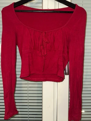 Röd croppad topp med knyt - Trendig röd croppad topp med långa ärmar och snygg knytning framtill. Toppen har en djupare rundad urringning och är tillverkad i ett mjukt, stretchigt material som sitter bekvämt. Perfekt att styla med höga jeans eller kjol.