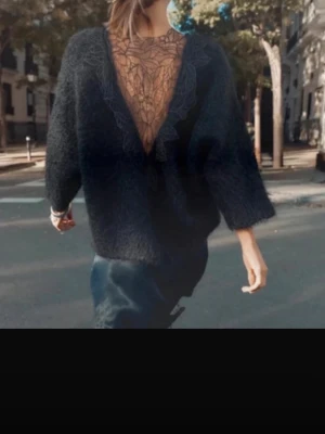 Svart stickad tröja med spetsrygg - Trendig svart stickad tröja med lös passform och trekvartsärmar. Baksidan har en djup V-ringning med transparent spetsdetalj och bladmönster som ger en unik look. Perfekt för dig som vill ha något extra till din outfit.