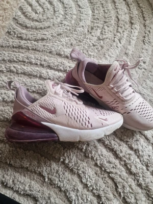 Nike Air Max 270 rosa sneakers - Säljer ett par Nike Air Max 270 sneakers i ljusrosa med mörkare rosa detaljer och vit sula. Skorna har en luftig mesh-överdel, synlig Air Max-dämpning i hälen och klassisk Nike-logga på sidan och plösen. Perfekta för dig som vill ha både stil och komfort.