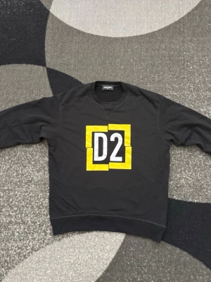 Dsquared2 tröja - Cool svart sweatshirt från Dsquared2 med stort D2-tryck i vitt och gult på bröstet. Tröjan har rund halsringning och ribbade muddar vid ärmslut och nederkant. Perfekt för dig som gillar streetwear och vill sticka ut med en snygg logga. Knappt använd STRLK 14Y
