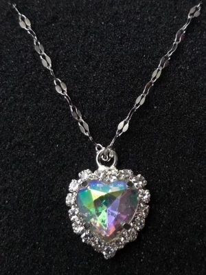 Halsband med regnbågshjärta och stenar - Snyggt halsband med ett hjärtformat hänge i regnbågsfärgad kristall, omgiven av gnistrande klara stenar. Kedjan är silverfärgad med dekorativa länkar. Perfekt för dig som gillar unika och glittriga smycken. Kedja i rostfritt stål 45 cm + 5 cm förlängningskedja.