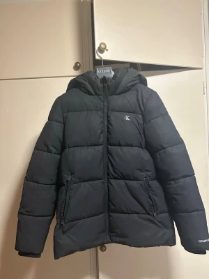 Svart pufferjacka från Calvin Klein - Svart pufferjacka från Calvin Klein Jeans med huva och dragkedja framtill. Jackan har två dragkedjeförsedda fickor och logga på bröstet samt ärmslutet och inuti jackan, säljer för för stor för mig.