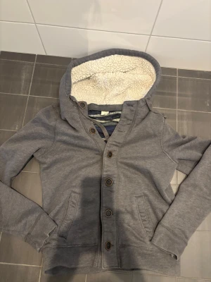 Grå hoodie från Hollister M - Mysig grå hoodie med knäppning framtill och två fickor från Hollister. Insidan av huvan är fodrad med vitt teddyfoder för extra värme och stil. Perfekt för kalla dagar och chill outfits. Klassisk passform och långa ärmar.