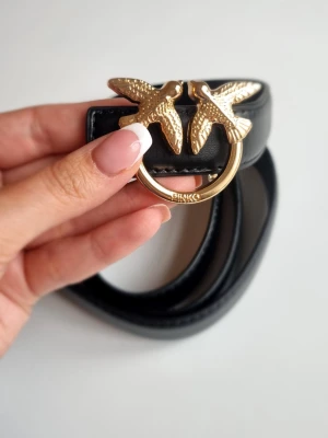 Svart skärp från Pinko med gulddetalj - Snyggt svart skärp från Pinko med ikoniskt spänne i guldformat metall med två fåglar. Skärpet är gjort i slätt skinn och passar perfekt för att lyfta vilken outfit som helst.